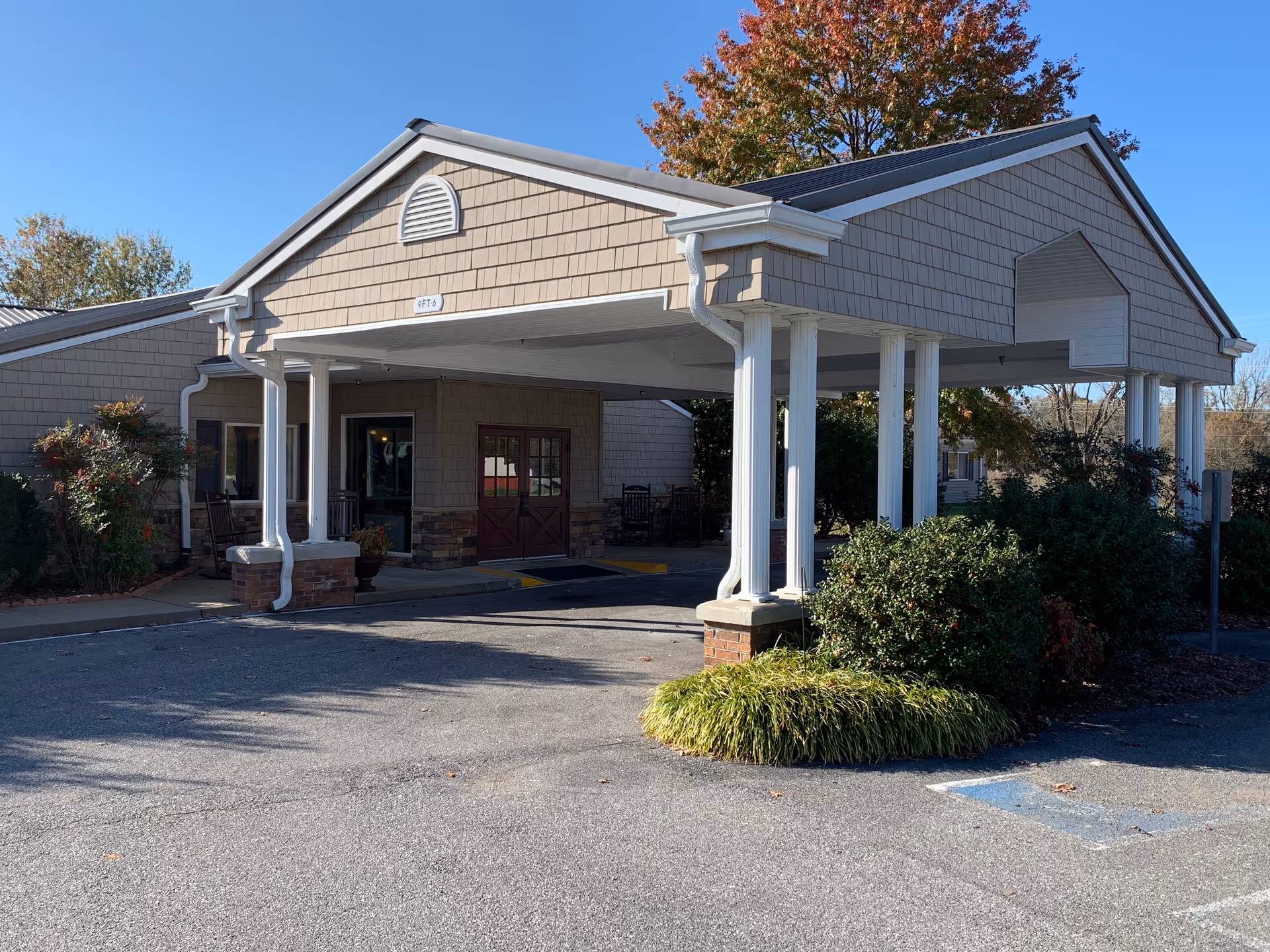 Entrance of a retirement community building with a covered drop-off area supported by white columns, surrounded by bushes and trees under a clear blue sky.