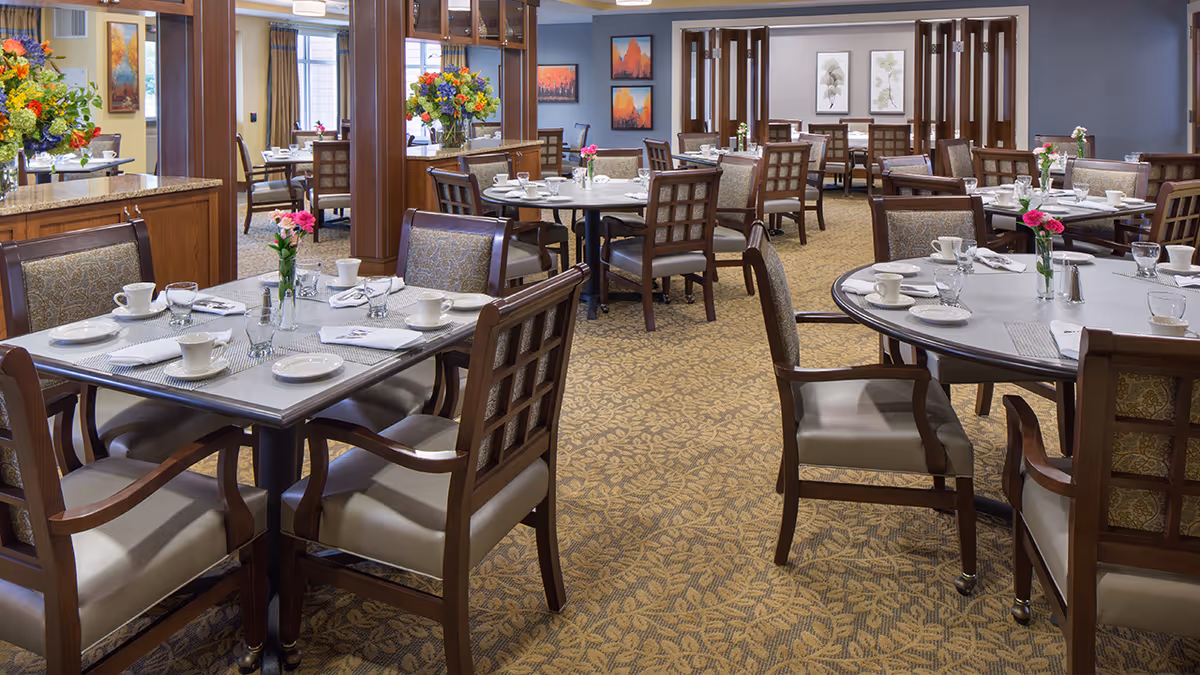 A well-lit dining room in a senior living facility with multiple tables set for a meal. Each table has chairs with cushioned seats and backs, white plates, cups, glasses, and napkins neatly arranged. There are small flower vases with pink flowers on each table. The room features carpeted flooring with a leaf pattern, wooden furniture, and colorful floral arrangements on a counter. Artwork is displayed on the walls, and large windows allow natural light to enter.