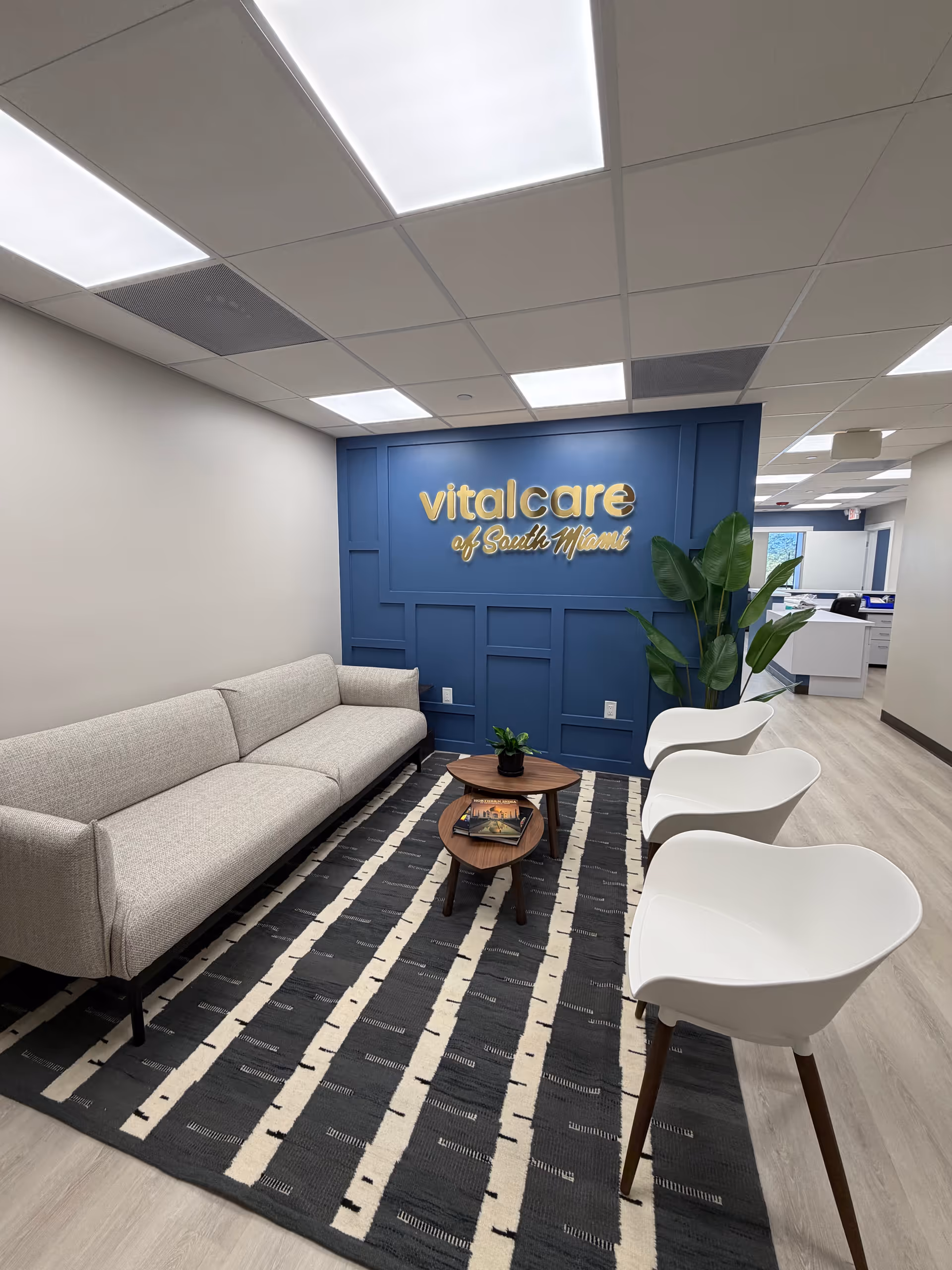 Lounge with gray sofa, white chairs, and blue wall sign reading Vital Care of South Miami.