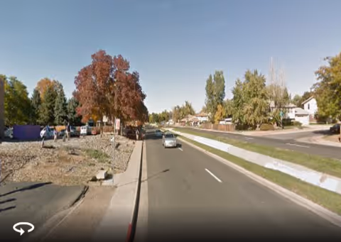 A suburban street scene with parked cars, trees, and houses along a sunny road.