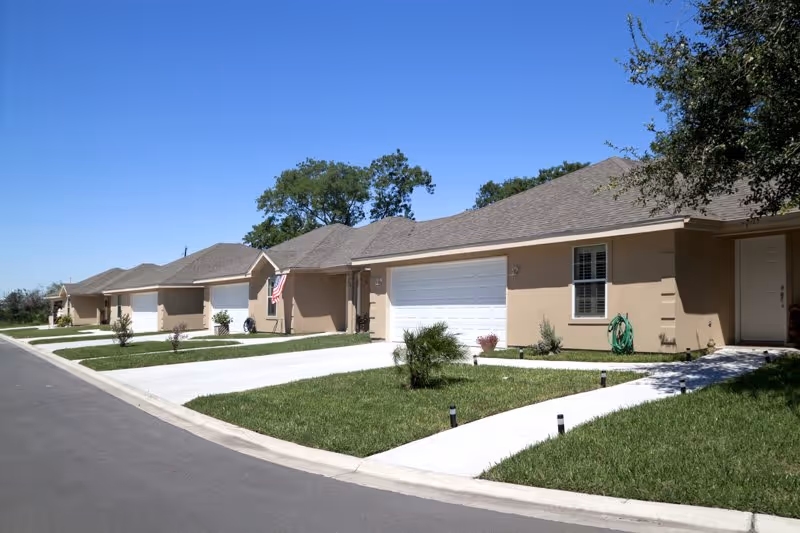 Row of beige single-story homes with garages and green lawns along a sunny street.