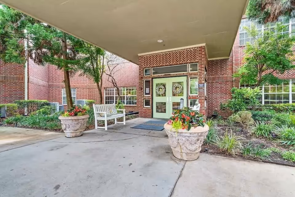 Entrance to a brick building with double green doors decorated with wreaths, flanked by large flower pots with colorful plants. There is a white bench to the left and greenery including trees and shrubs surrounding the entrance area.