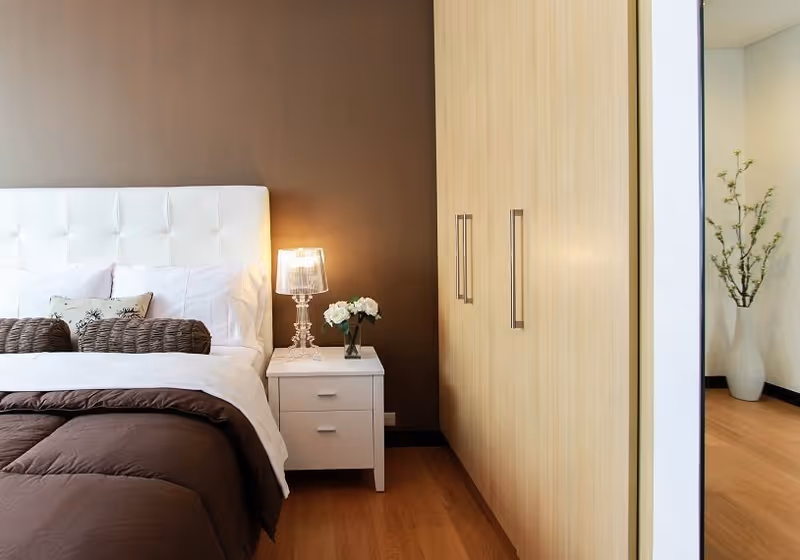 A modern bedroom with a white tufted headboard, nightstand and lamp, brown bedding, and a built-in light wood wardrobe.
