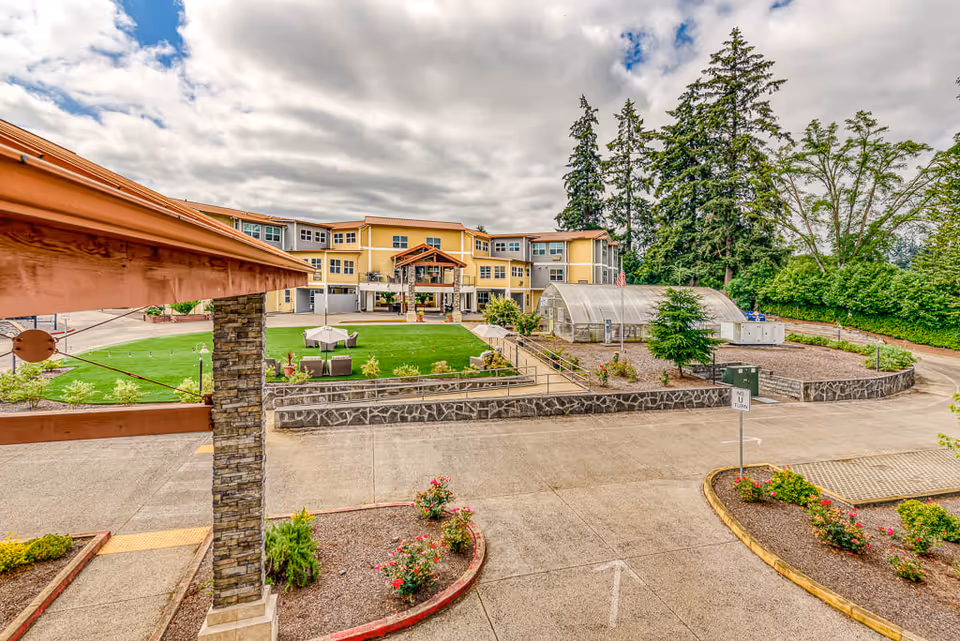 View of a senior living facility courtyard and the front of a three-story yellow building with a greenhouse, lawn seating, and driveway.