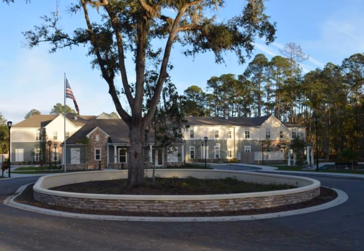 Two-story senior living building seen from a circular driveway with a landscaped island and large tree, an American flag, and trees in the background.