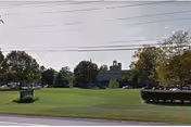 Grassy grounds with trees and a distant facility building visible beyond a road.