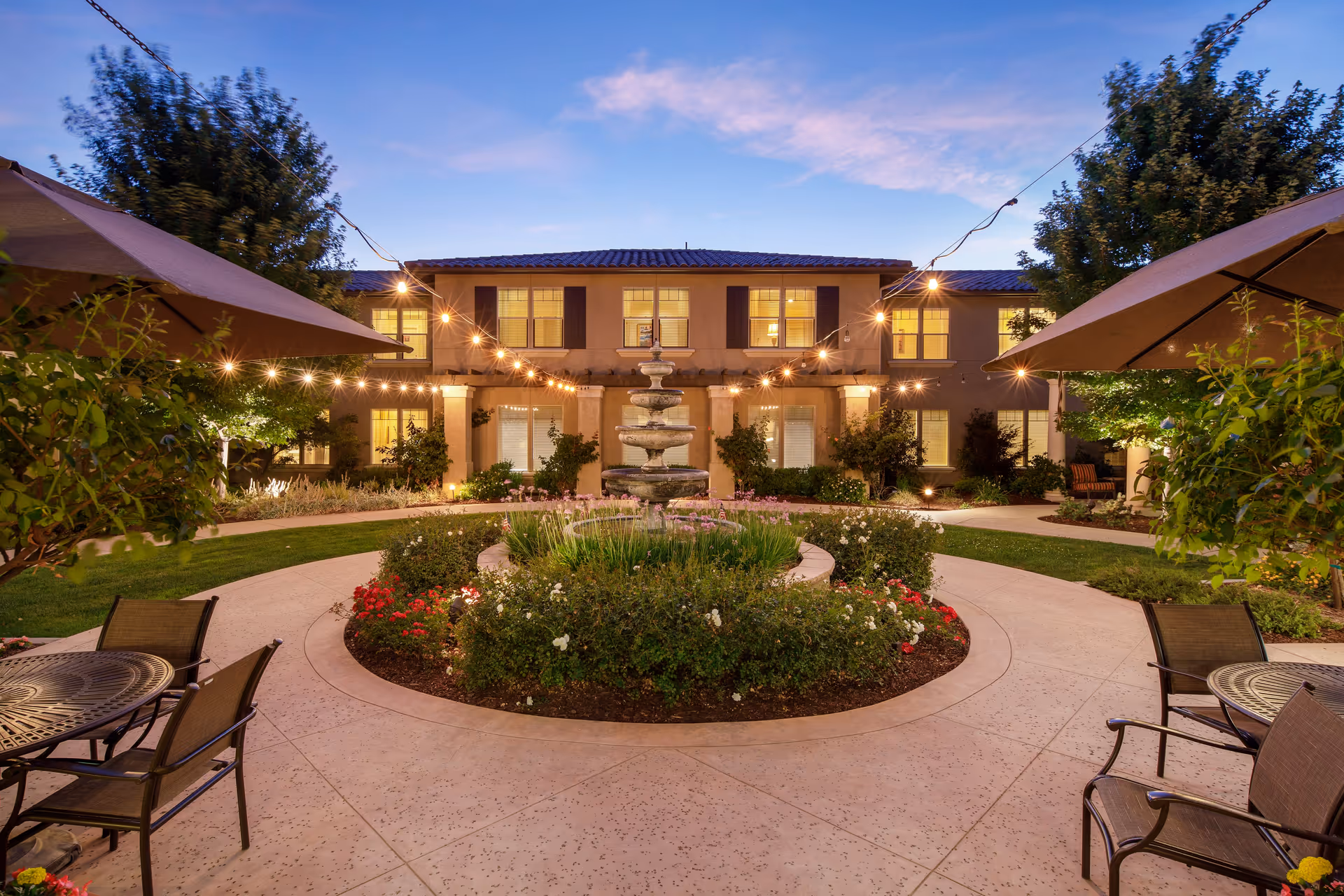 Outdoor courtyard at dusk with a central circular garden featuring a multi-tiered fountain surrounded by flowers and greenery. String lights are hung above, creating a warm ambiance. There are patio tables with umbrellas and chairs on either side of the courtyard. The two-story building in the background has lit windows and a tiled roof.