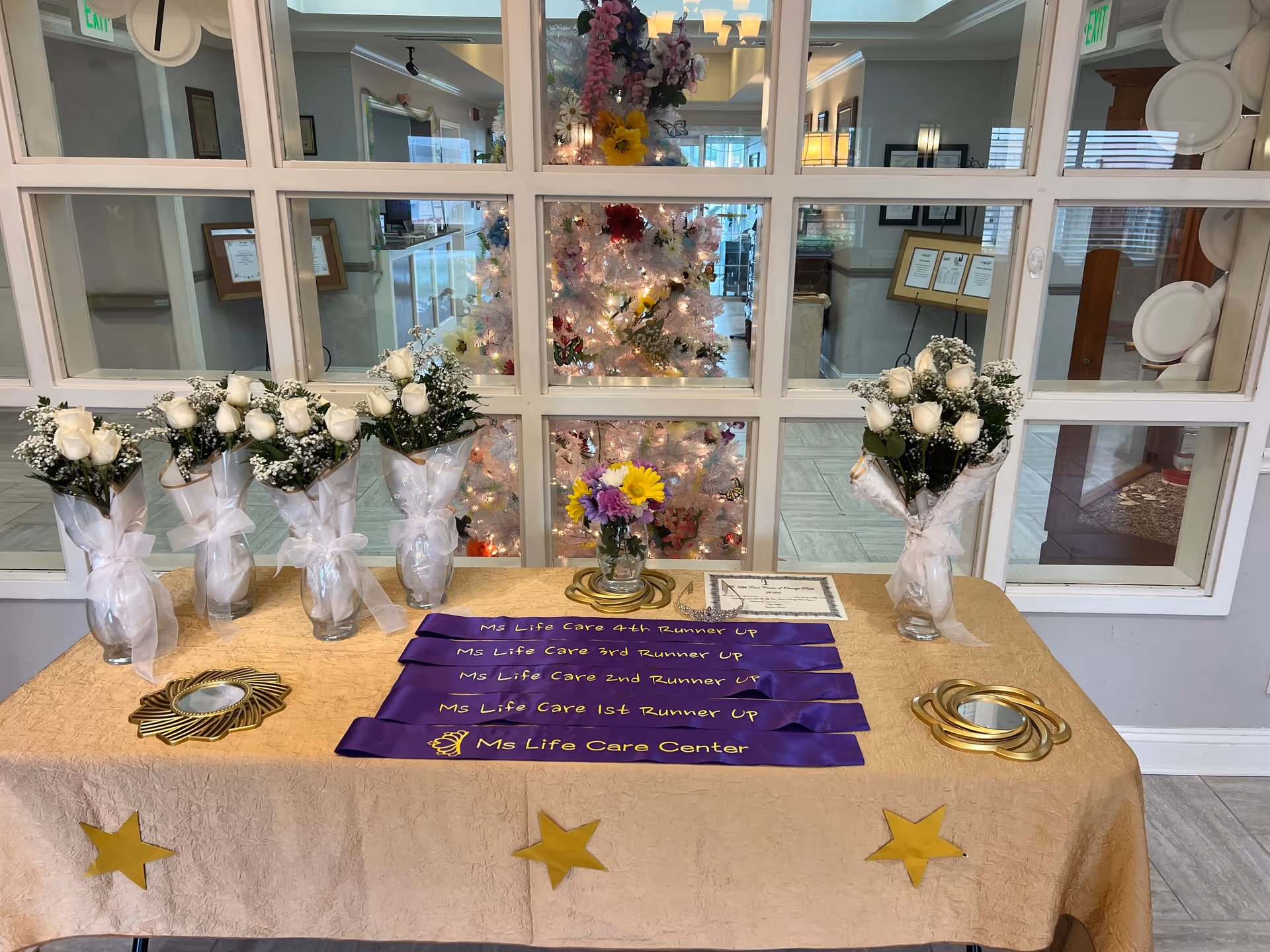 Lobby display table with white flower bouquets and purple ribbons; glass walls reveal a Christmas tree.