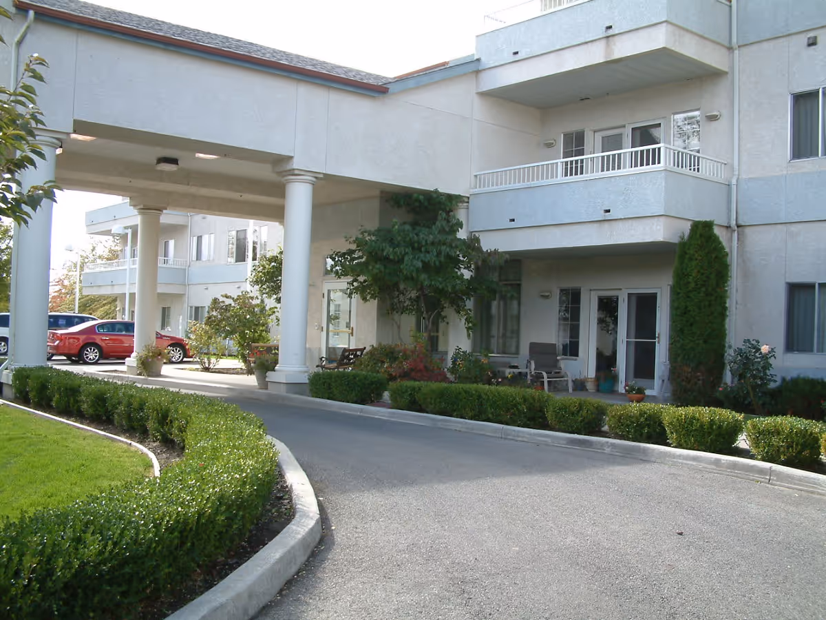 Exterior view of Wedgewood Park Senior Living showing a driveway with a covered entrance supported by white columns, landscaped bushes and trees, and a multi-story building with balconies and windows.