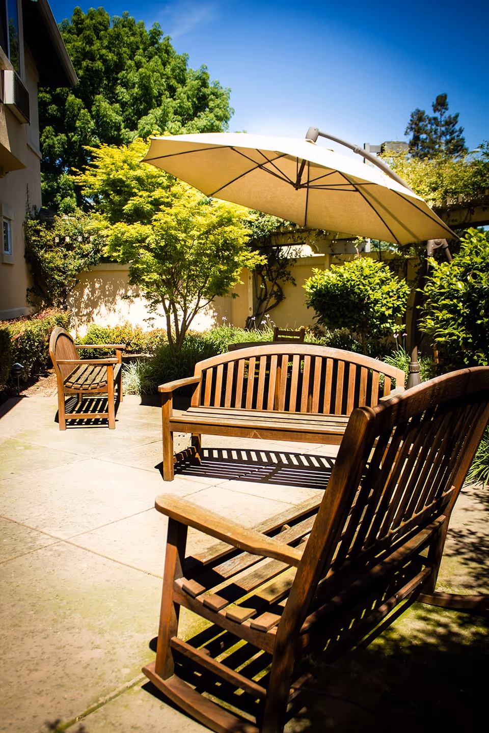 A sunny outdoor area with wooden benches and an umbrella surrounded by greenery.