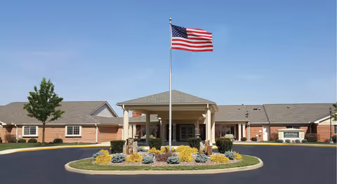 Front entrance of a single-story senior living building with a circular driveway, landscaped island and a central flagpole flying the American flag.