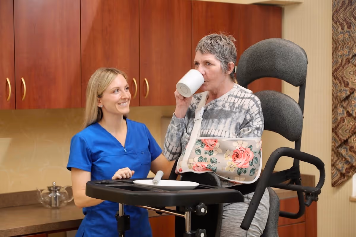 A healthcare worker in blue scrubs smiling and assisting an elderly woman with a floral arm sling who is drinking from a white mug while seated in a supportive chair with a tray table in a room with wooden cabinets.