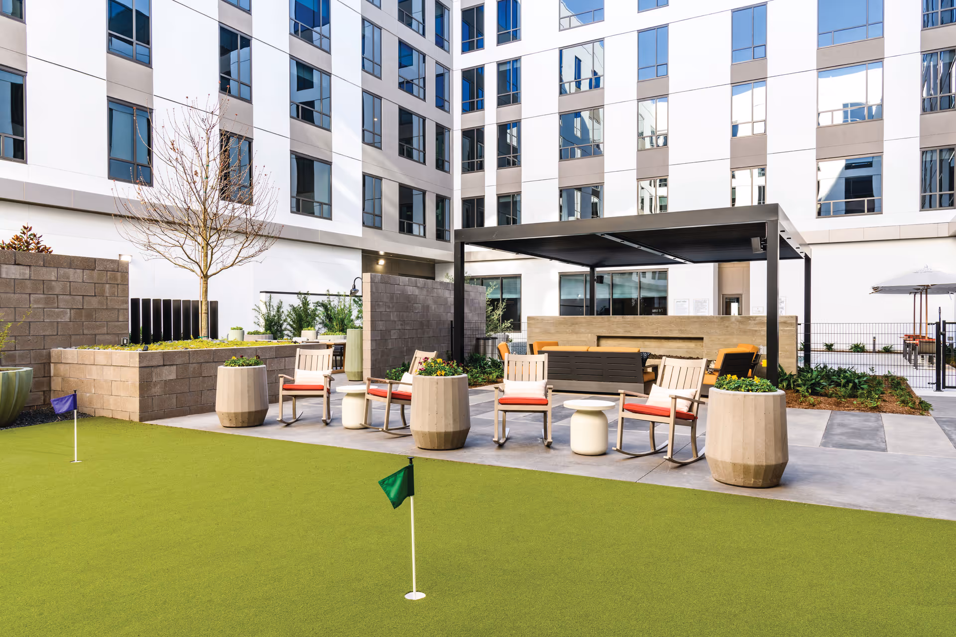 Outdoor courtyard area at Clearwater at The Heights featuring a putting green with small flags, several chairs with red cushions arranged around small tables, large planters with greenery, and a shaded seating area under a modern pergola. The courtyard is surrounded by a multi-story building with many windows.