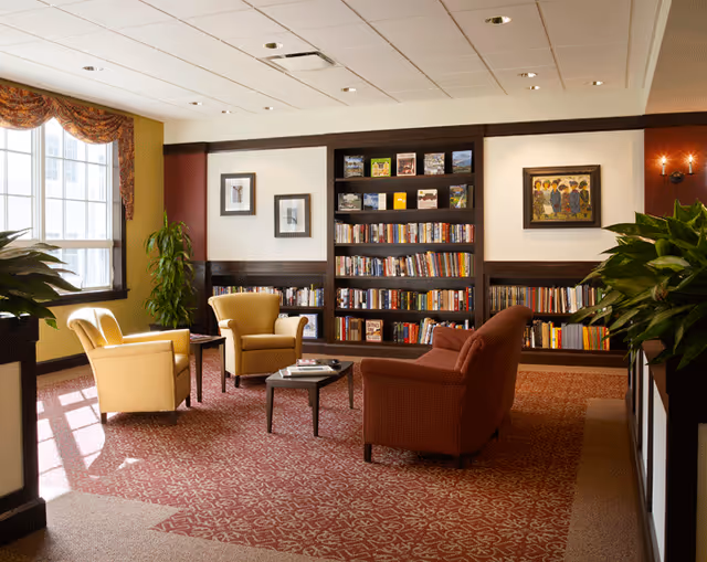 A cozy reading lounge with a large bookshelf filled with books and magazines, three comfortable armchairs arranged around a small coffee table, large windows with decorative curtains letting in natural light, framed artwork on the walls, and potted plants adding greenery to the space.