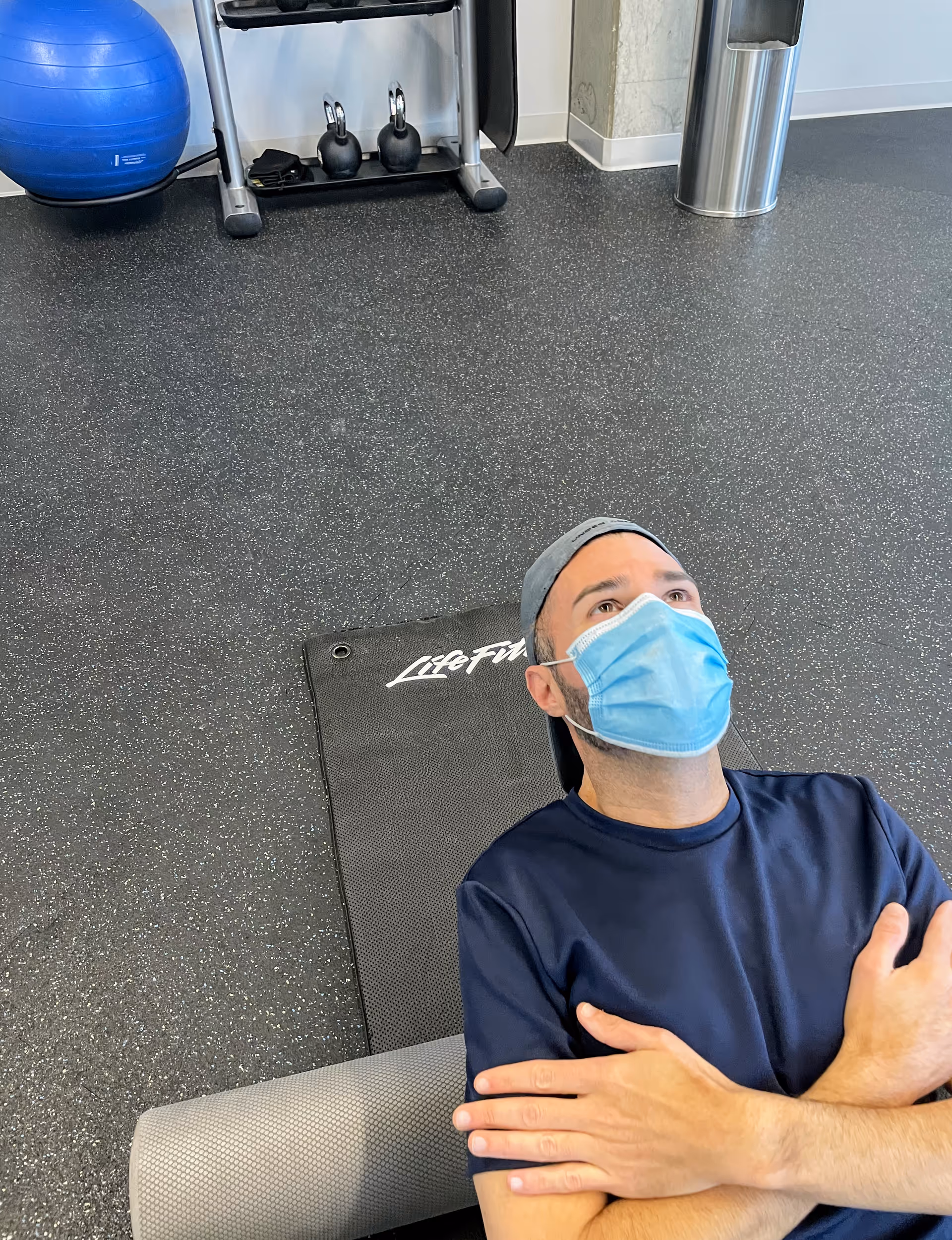 A man wearing a blue mask lies on a workout mat in a gym.