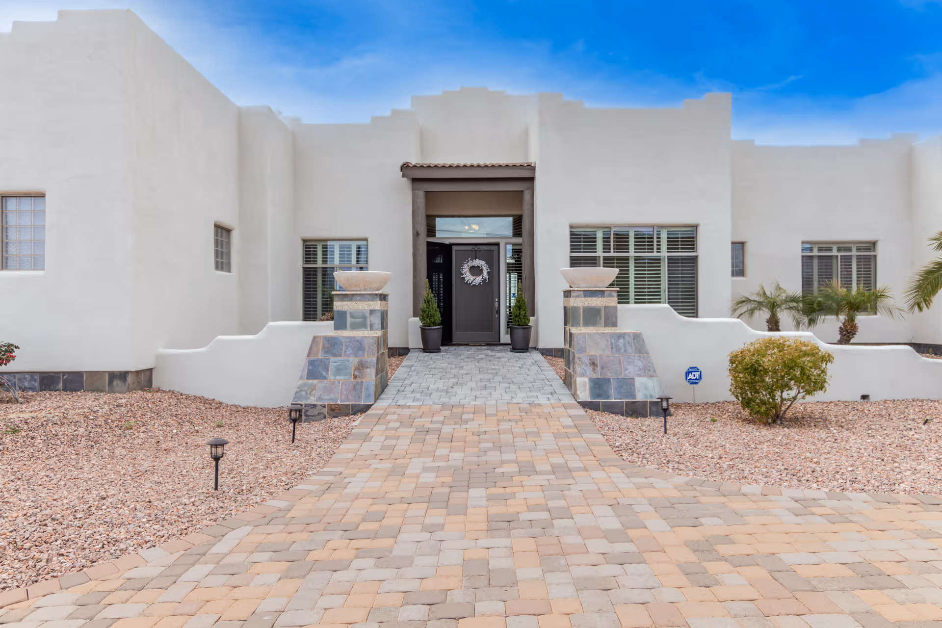 White stucco building entrance with a paved walkway and desert landscaping leading to a central doorway.
