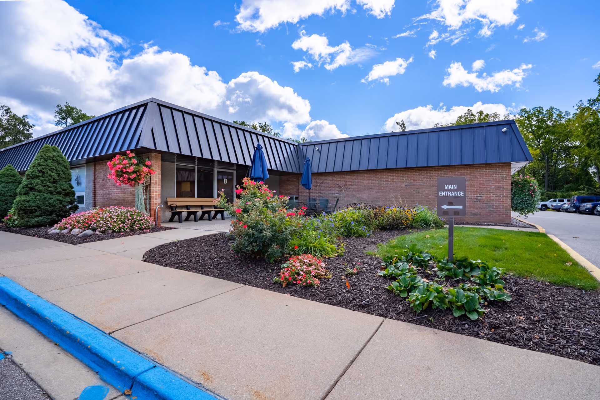 Exterior view of a single-story brick building with a dark metal roof, surrounded by landscaped flower beds and shrubs. There is a sidewalk and a sign indicating the main entrance with an arrow pointing to the right. The sky is partly cloudy with blue patches.