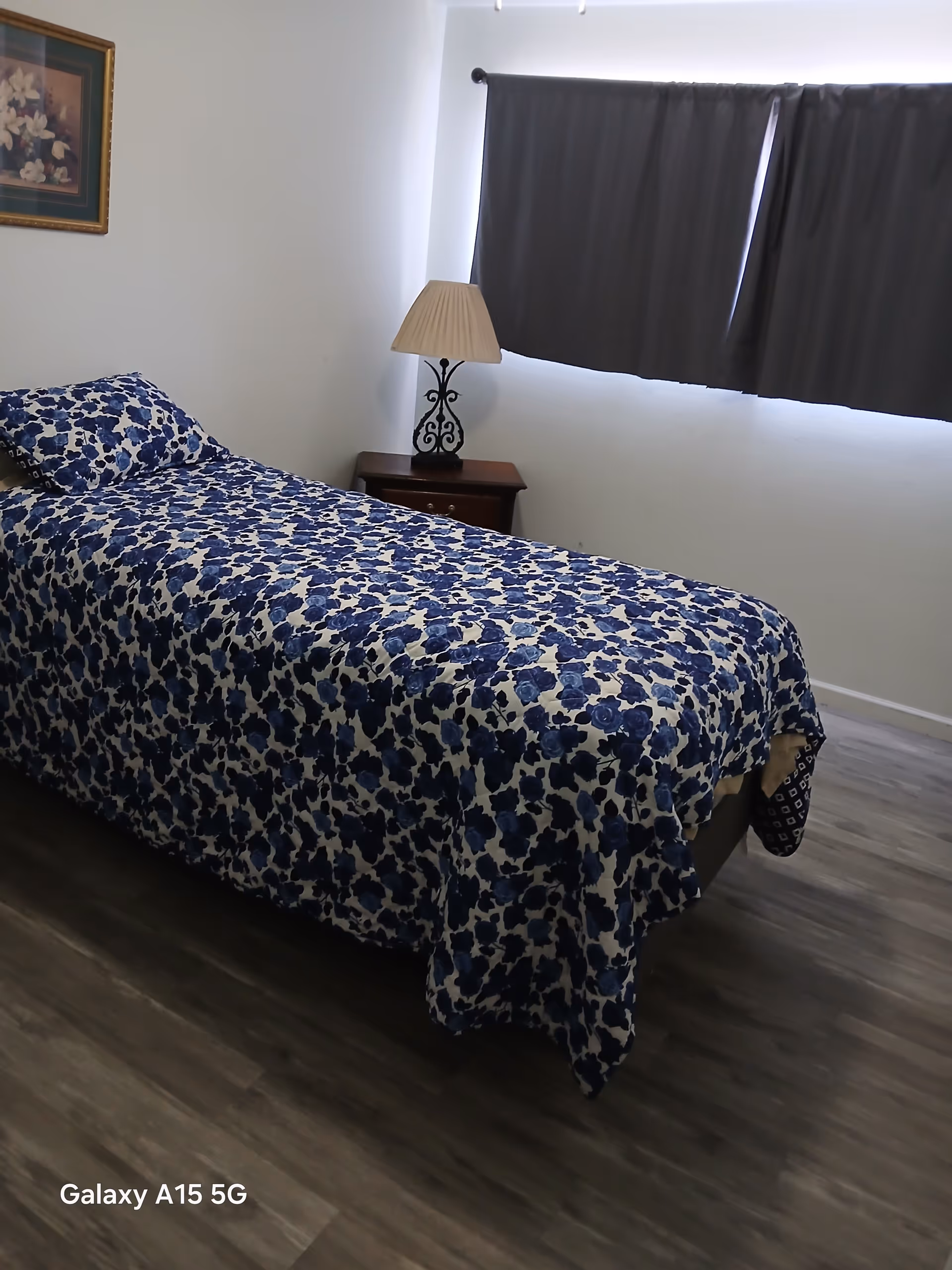 Bedroom with single bed, blue floral bedding, nightstand, lamp, and dark curtains.