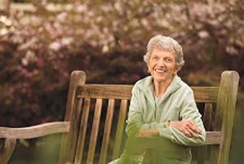 Smiling elderly woman sits on a wooden bench in an outdoor garden setting.