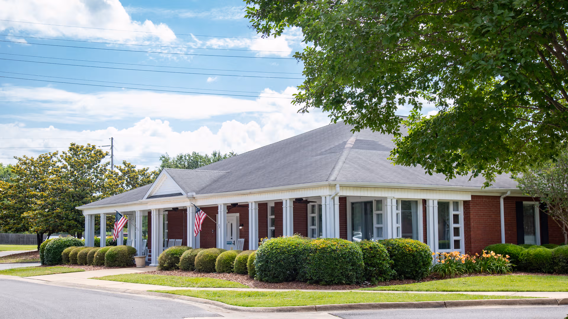 Single-story brick senior living facility with a front porch, shrubs, and flags.