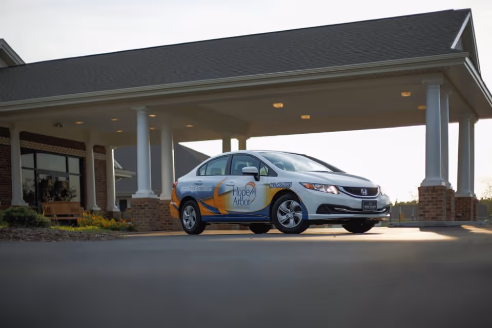 A white Honda sedan with Hope Arbor branding parked under a covered entrance of a brick building, likely the facility entrance.