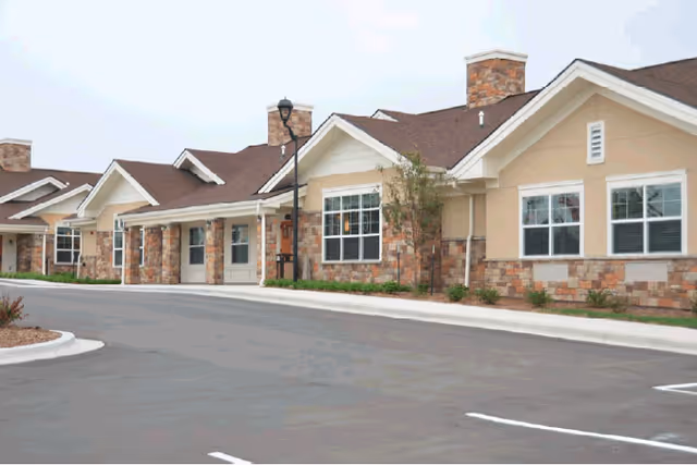 Exterior view of a senior living facility with brick and beige siding, parking lot in foreground.