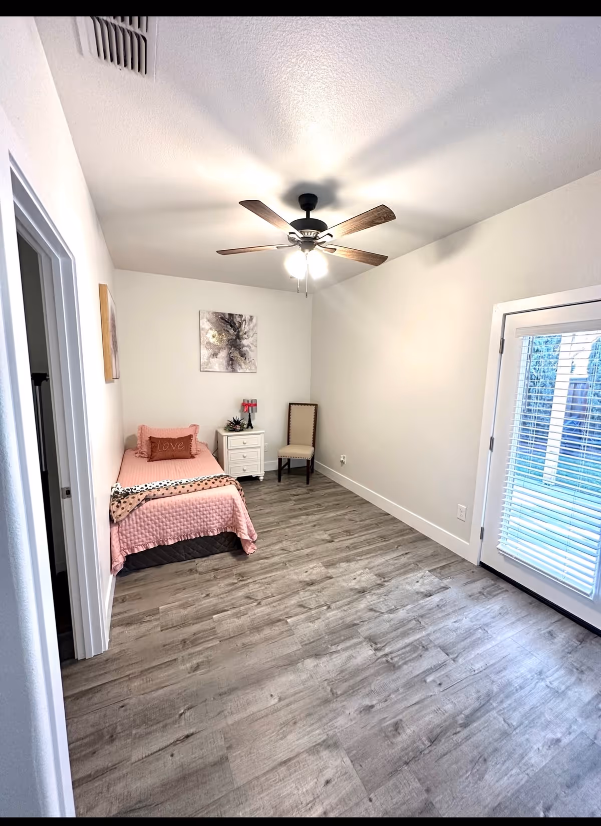 Small bedroom with pink bed, nightstand, chair, ceiling fan, and a door with blinds.