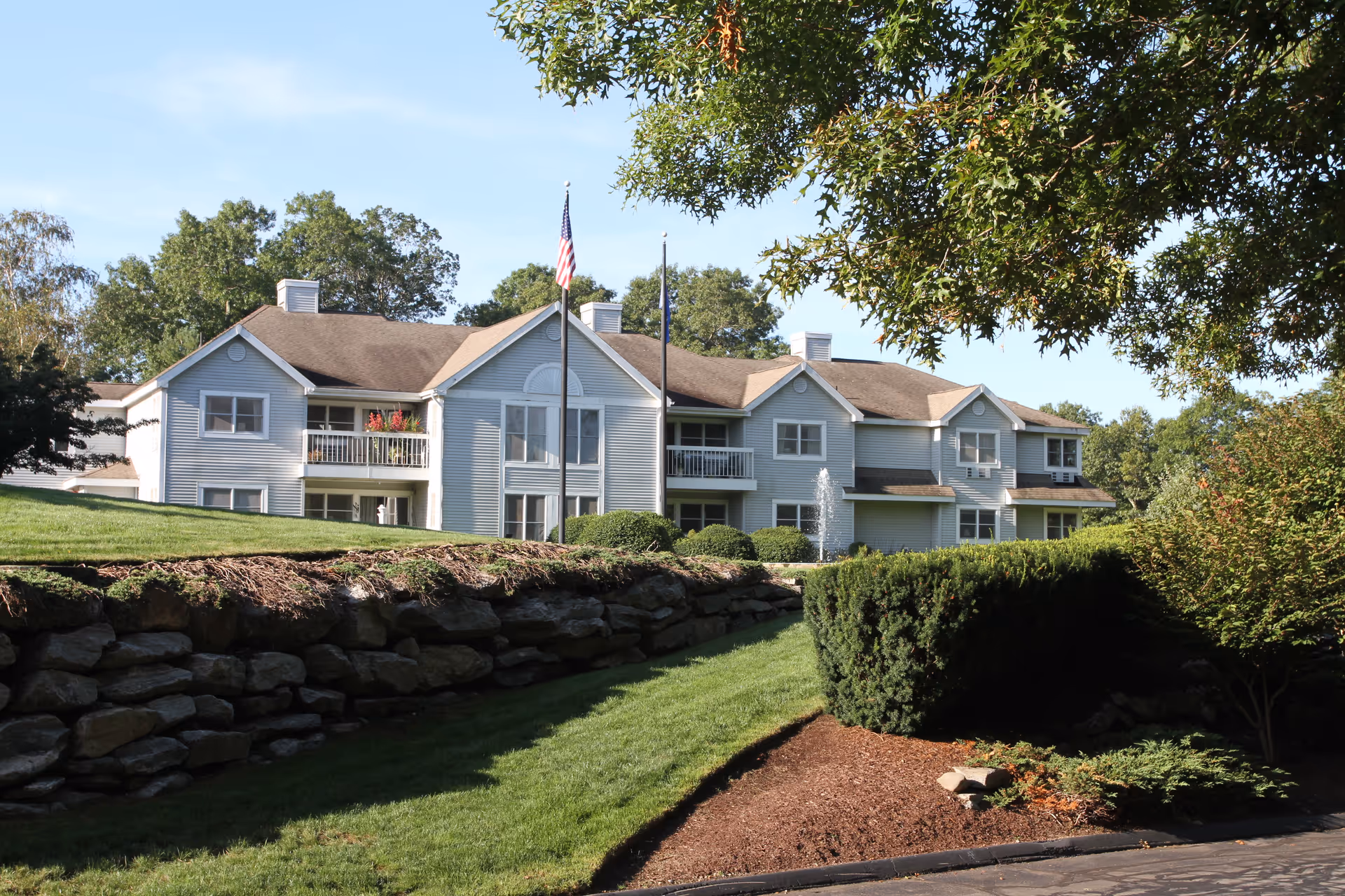 Exterior view of a two-story residential building with light gray siding and multiple balconies, surrounded by green lawns, bushes, and trees under a clear blue sky. Two flagpoles with flags are positioned in front of the building.