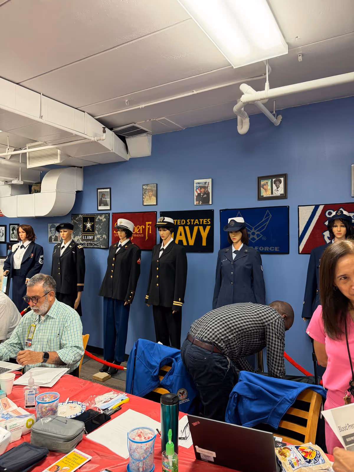 Indoor hall with uniformed mannequins lined along a blue wall and people seated.