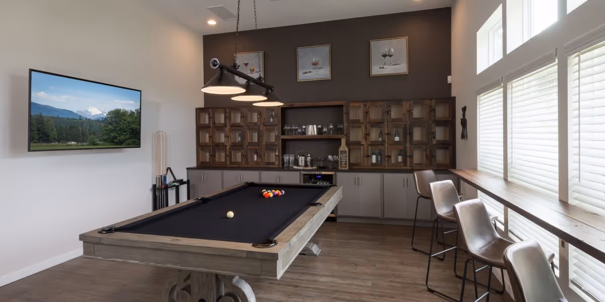 Recreation room with a pool table at center, wall-mounted TV, bar cabinets, and high windows with counter seating.