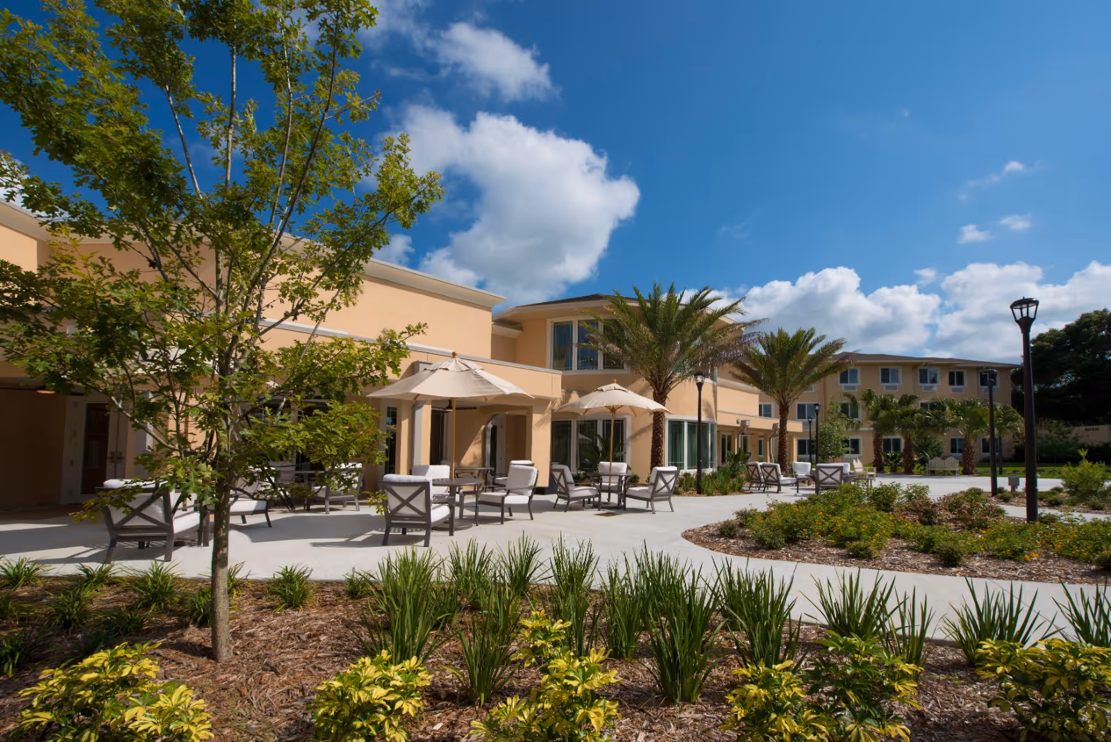 Outdoor patio area of a senior living facility with beige buildings, palm trees, green shrubs, and several seating arrangements with umbrellas under a blue sky with some clouds.