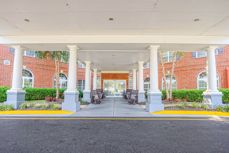 Covered entrance area of a brick building with white columns and rocking chairs arranged on both sides of the walkway leading to glass double doors. There are small landscaped areas with bushes and trees on either side of the entrance.