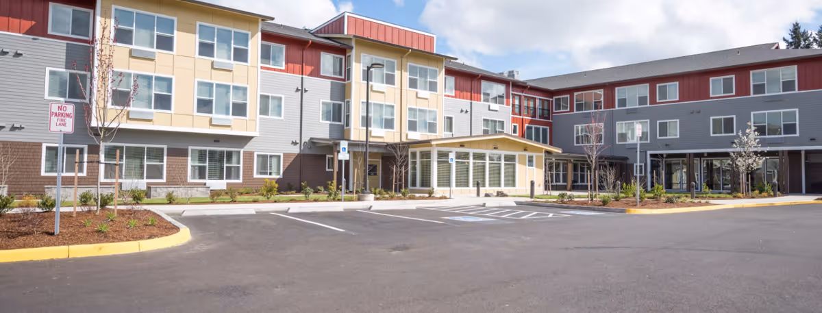 Exterior view of a modern three-story senior living facility with a large parking area in front, landscaped with small trees and shrubs, under a partly cloudy sky.