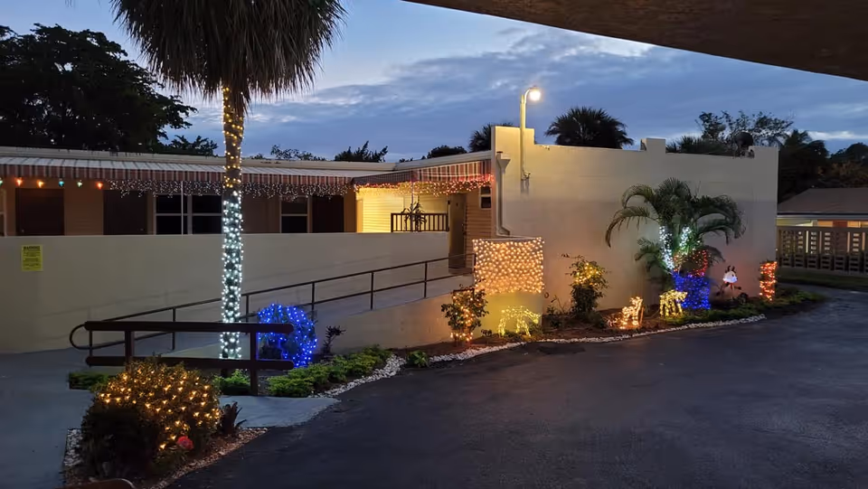 Dusk view of a senior living building entrance with a ramp, a palm tree and holiday lights and illuminated decorations along the driveway.