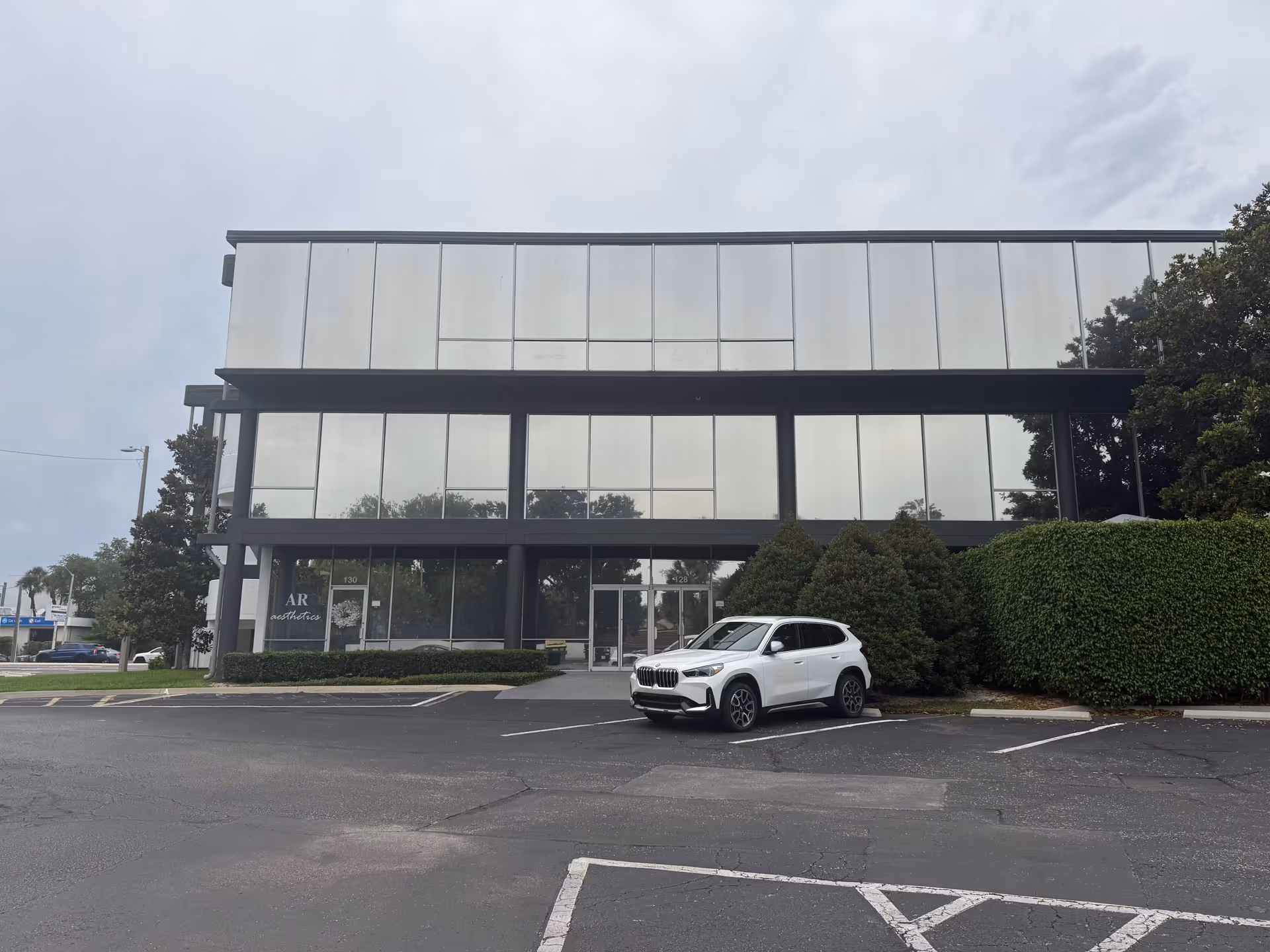 Modern two-story glass-front building with a parking lot and a white SUV in front.
