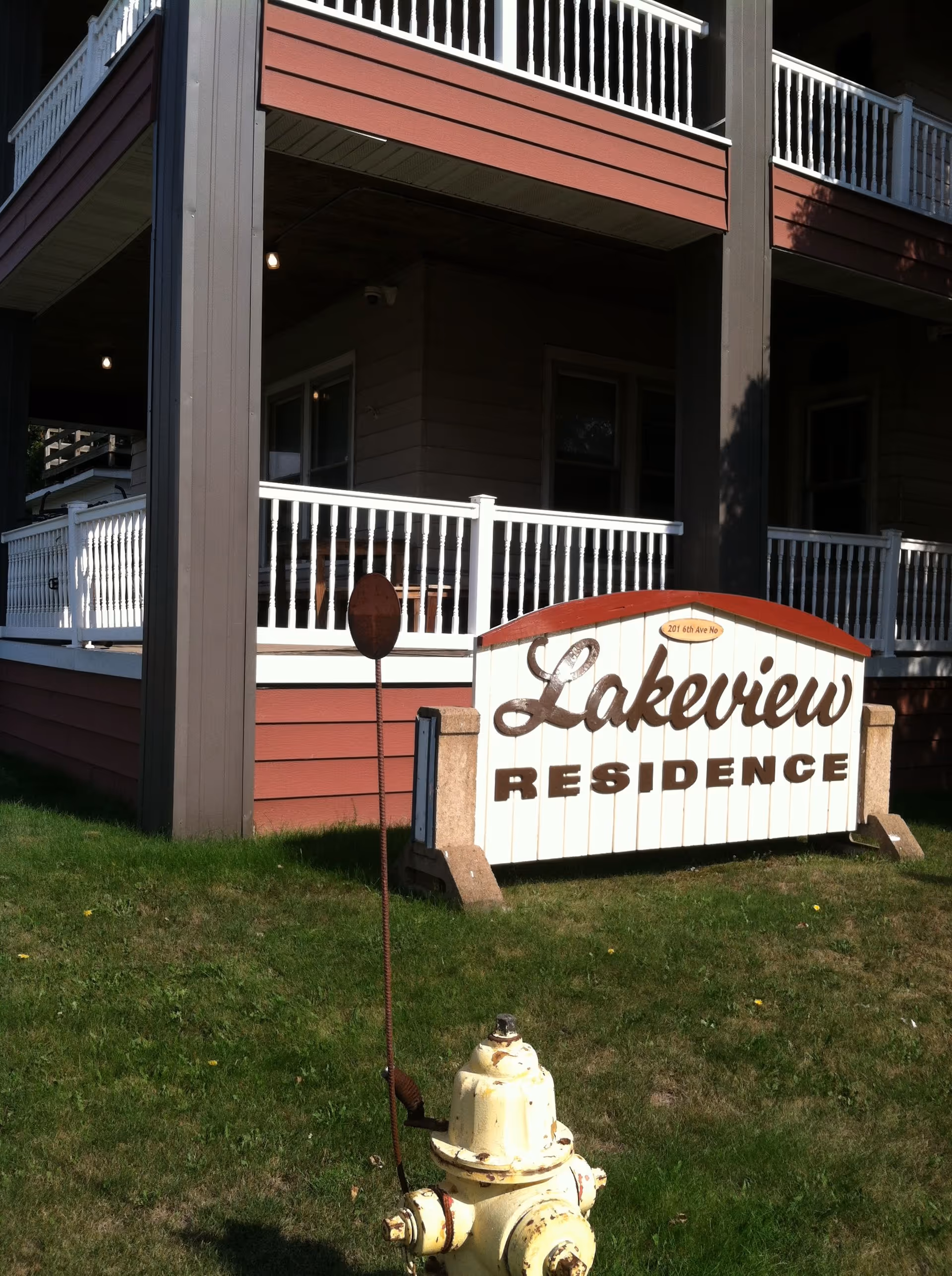 Exterior view of a building with white railings and red siding, featuring a sign that reads 'Lakeview Residence' on a grassy area in front. A yellow fire hydrant is visible in the foreground.