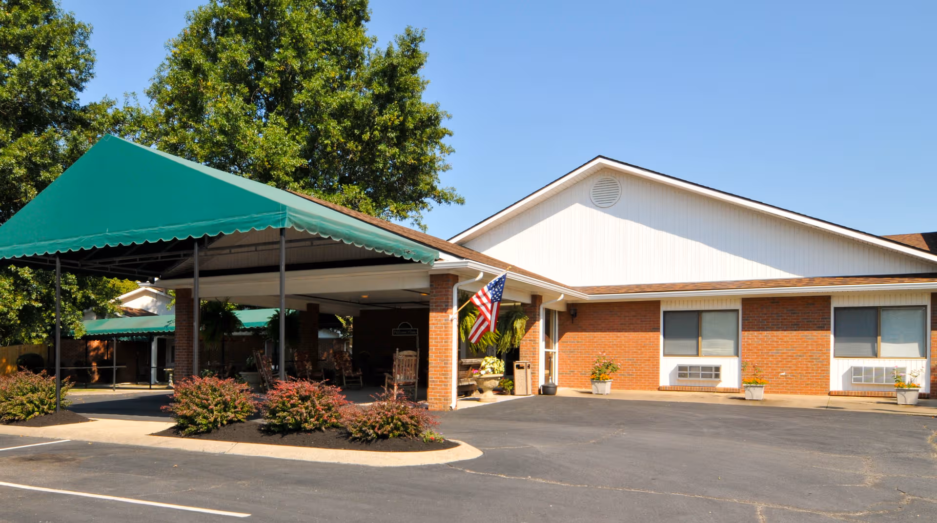 Brick senior living facility front with green canopies, a parking lot, and American flag at entrance.