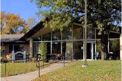 Front exterior of New Glen Oaks Nursing Home with glass facade, benches, and autumn trees.