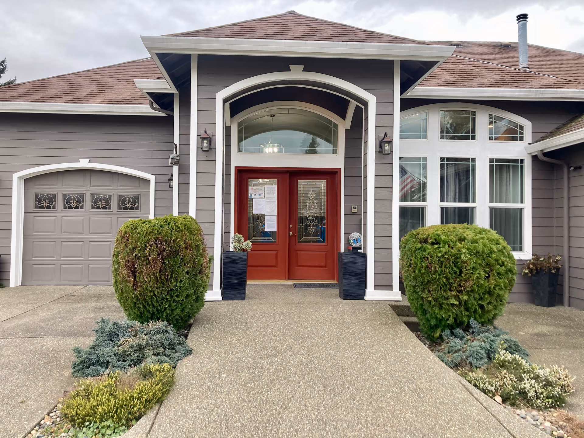 Gray senior living facility exterior with red double doors and trimmed shrubs at entrance.