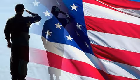 A silhouetted soldier salutes beside a large American flag outdoors.