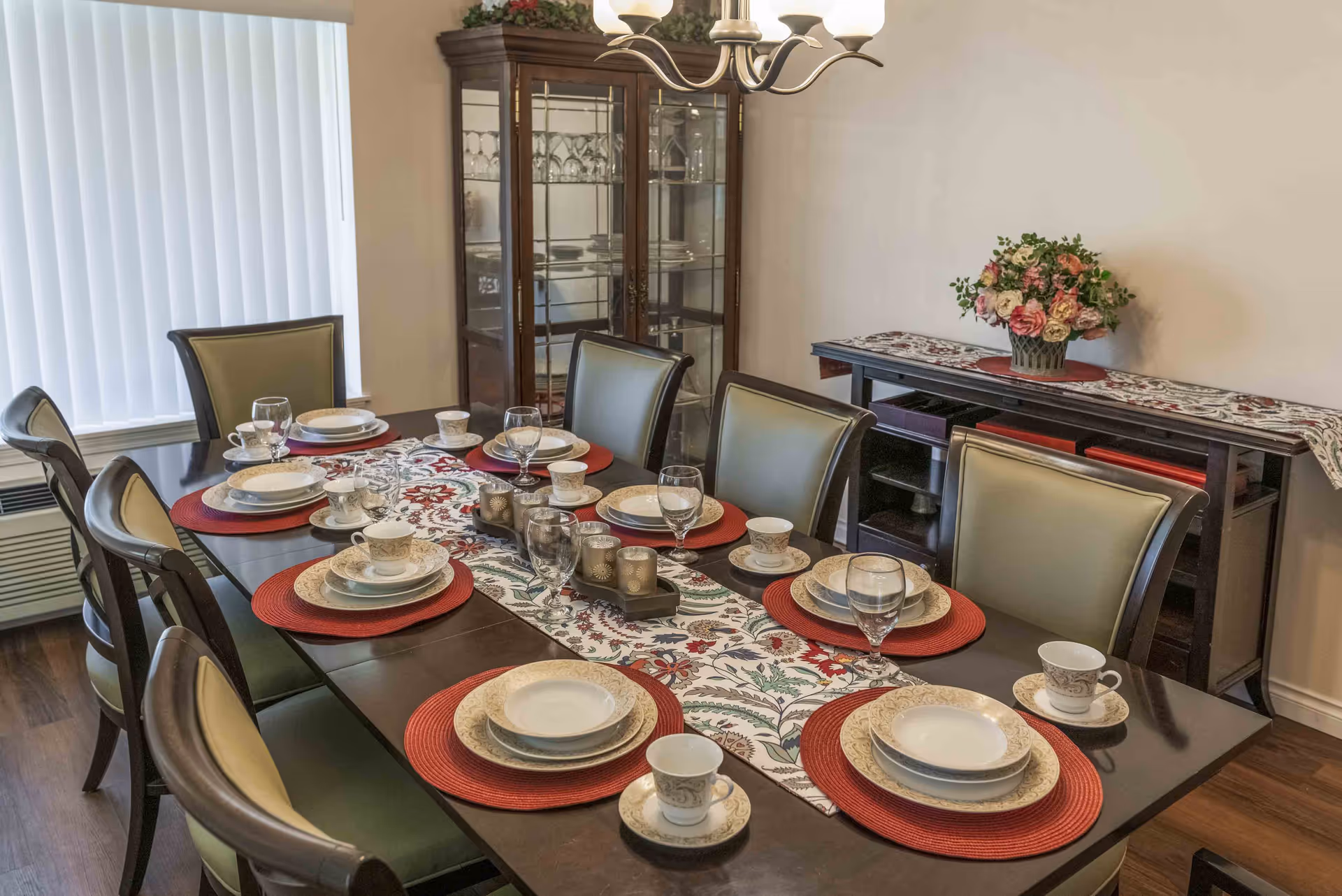 A dining room with a dark wooden table set for eight people. Each place setting includes a red placemat, a set of beige patterned plates, a teacup with saucer, and a water glass. A floral table runner runs down the center of the table with decorative candle holders. In the background, there is a glass-fronted china cabinet with glassware and plates, and a sideboard with a floral arrangement on top. The room has light-colored walls and a window with vertical blinds.