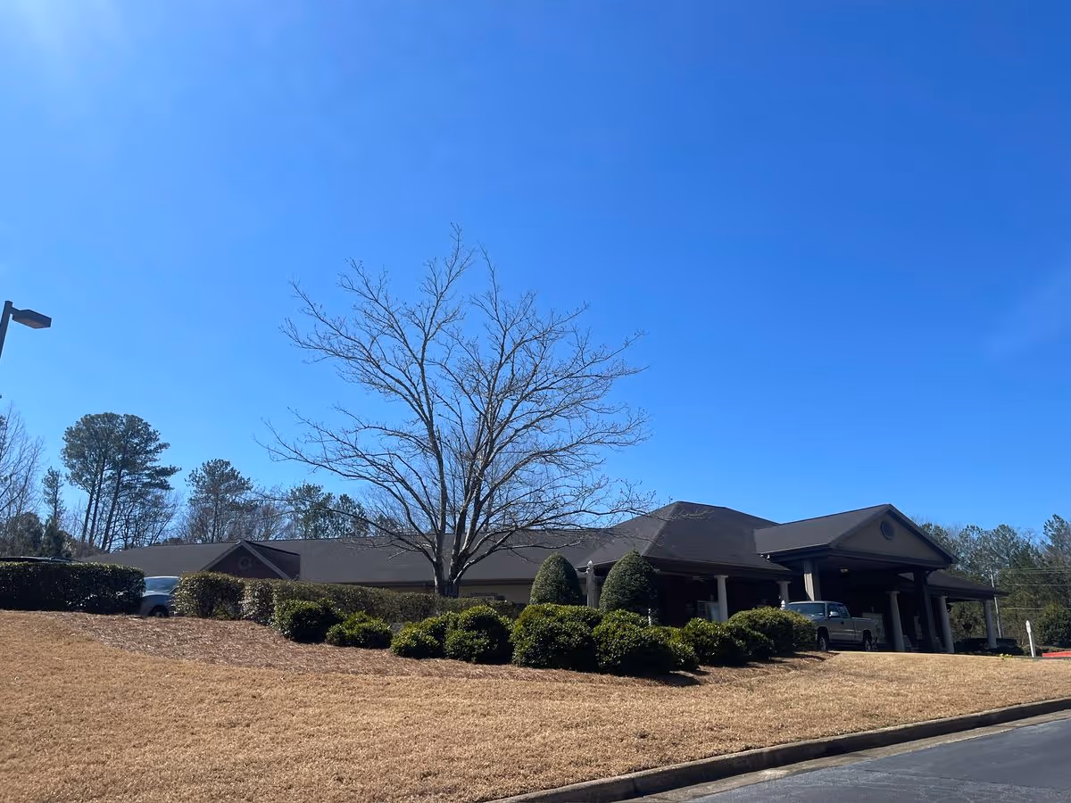 Single-story building with a covered portico entrance, a leafless tree and trimmed shrubs on a grassy slope under a clear blue sky.