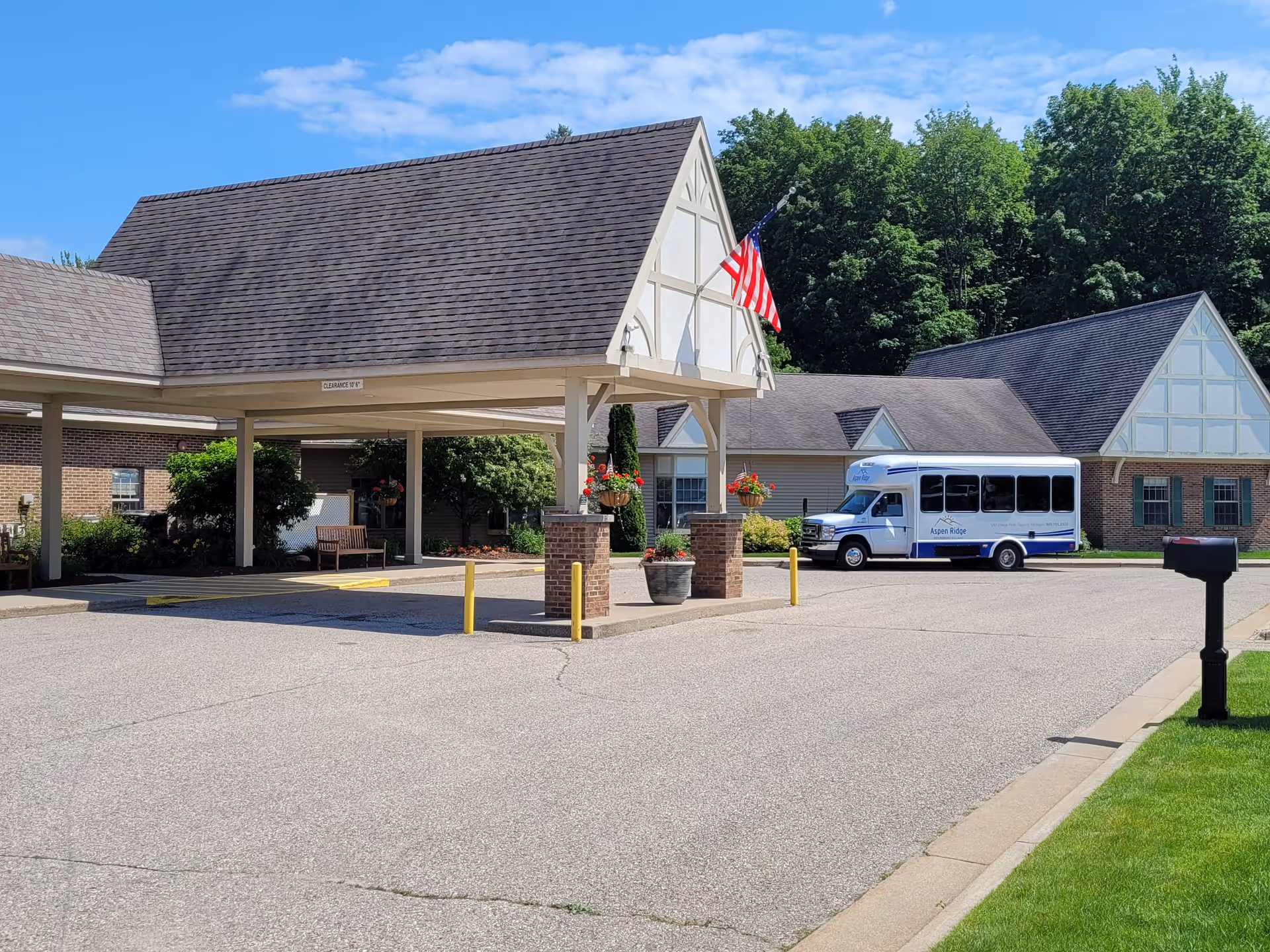 Exterior view of Aspen Ridge senior living facility showing a covered entrance with brick pillars and hanging flower baskets, an American flag, a white shuttle bus parked nearby, and trees in the background under a partly cloudy sky.