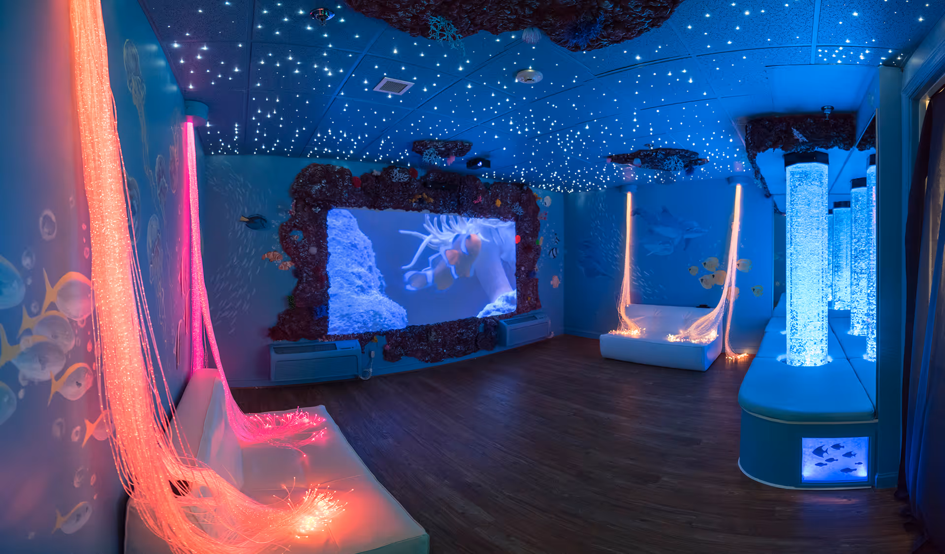 A sensory room with an underwater theme featuring blue walls decorated with fish and coral murals, fiber optic lights emitting pink and blue glows, bubble tubes, a ceiling with star-like lights, and a large screen displaying an underwater scene with clownfish.