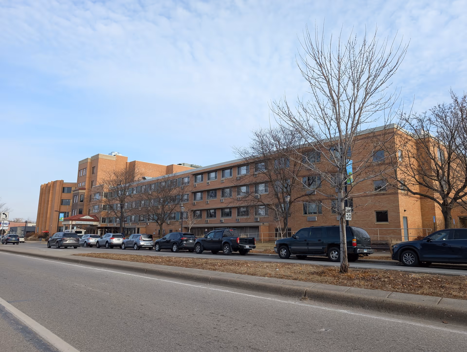 Long brick care facility building front along a city street with parked cars and leafless trees under a blue sky.