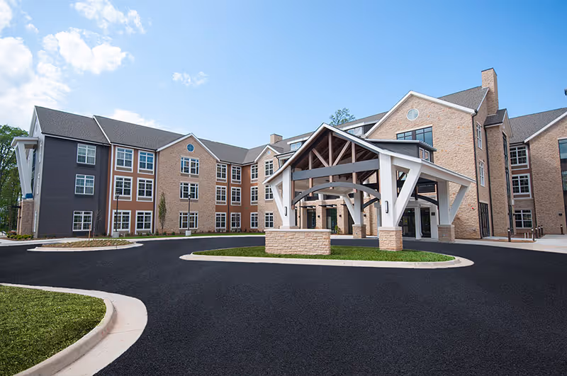 Front entrance of a multi-story brick senior living building with a covered porte-cochère and circular driveway under a blue sky.