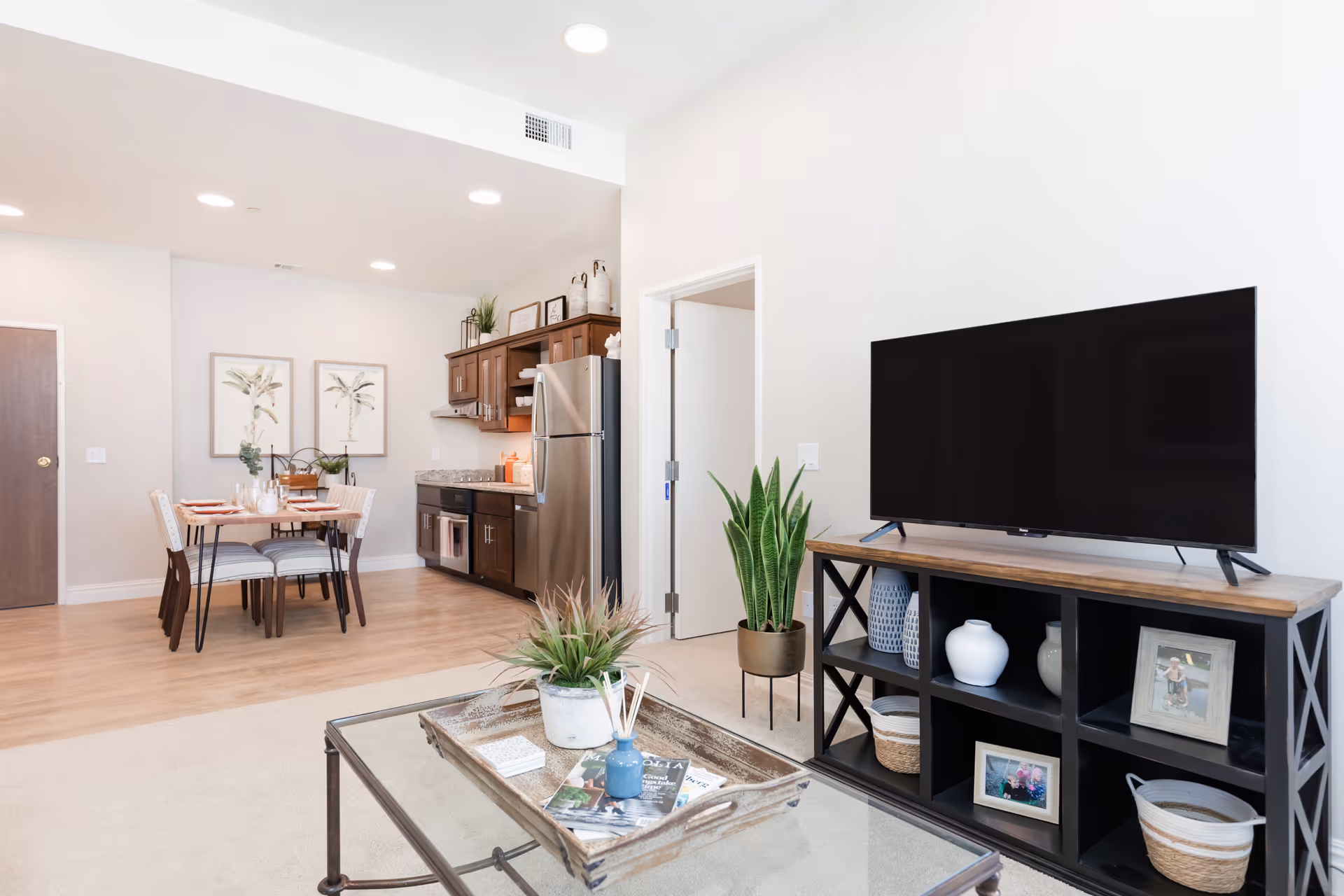 Bright and modern living area with a large flat-screen TV on a black wooden stand decorated with vases, baskets, and framed photos. In the foreground, a glass coffee table holds a tray with a plant and magazines. The background shows a dining area with a wooden table set for four and a kitchen with stainless steel appliances and dark wood cabinets.