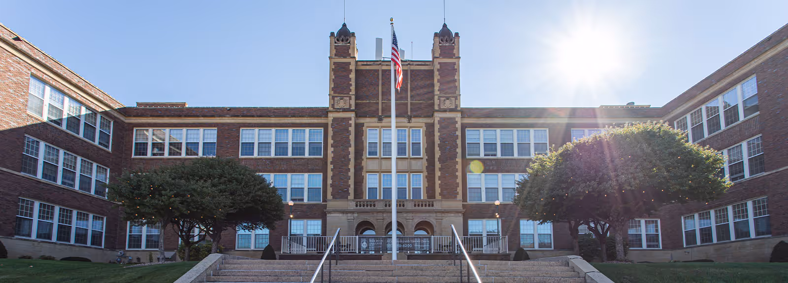 Front exterior of a three-story brick building with a central tower, flagpole, and steps leading to the main entrance under bright sunlight.