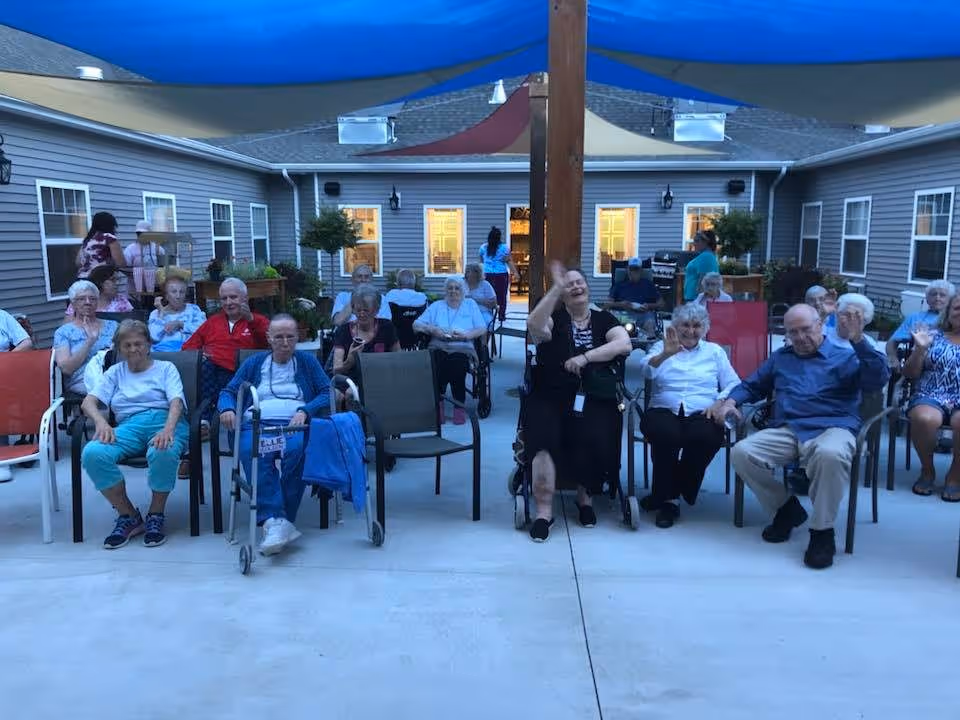 A group of elderly people sitting on chairs and wheelchairs in an outdoor courtyard area of a retirement living facility during the evening. The courtyard is surrounded by a single-story building with windows and doors, and there are shade sails overhead. Some people are waving and smiling at the camera.