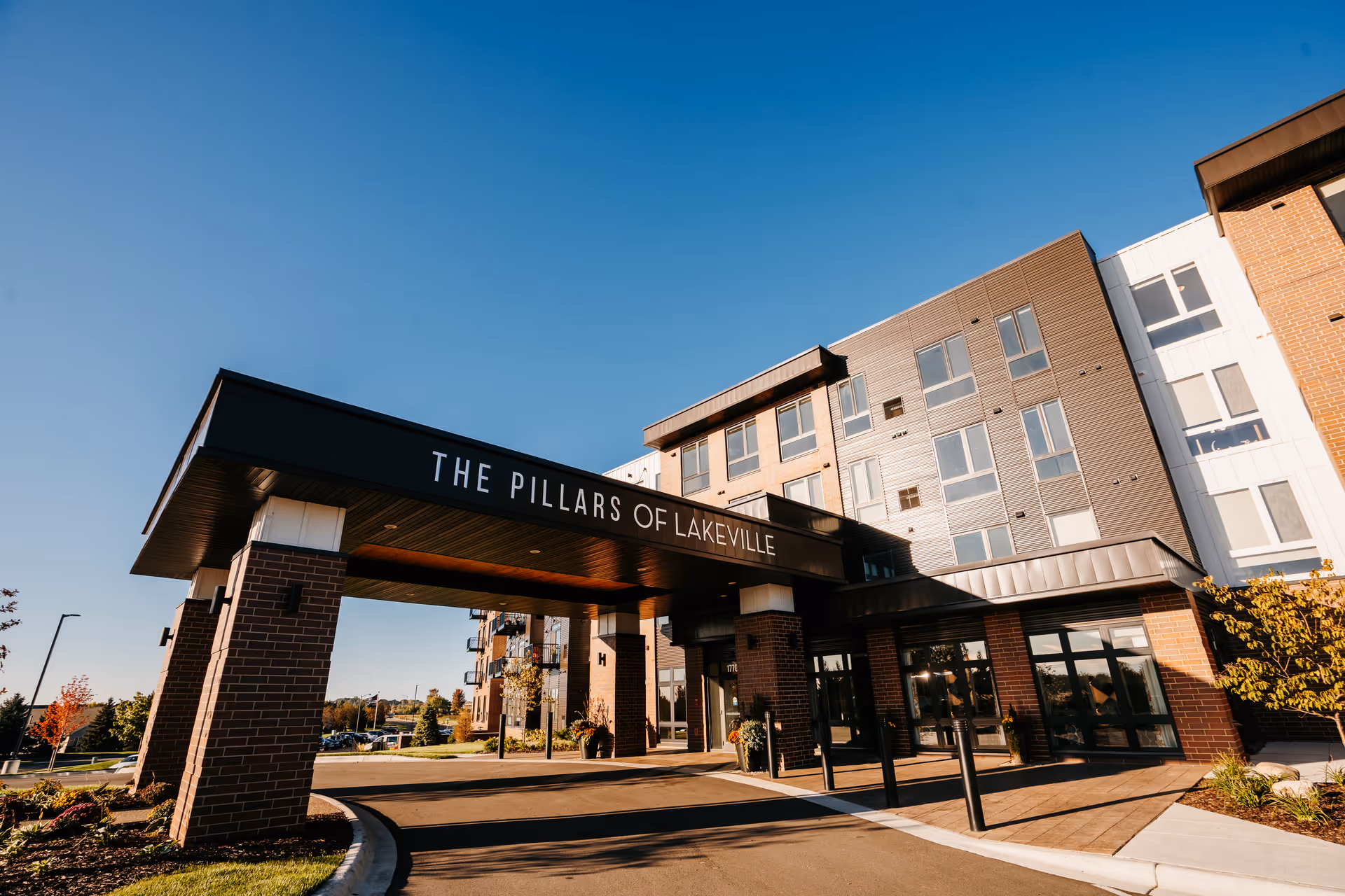 Exterior view of The Pillars of Lakeville building entrance with a covered driveway supported by brick pillars under a clear blue sky.